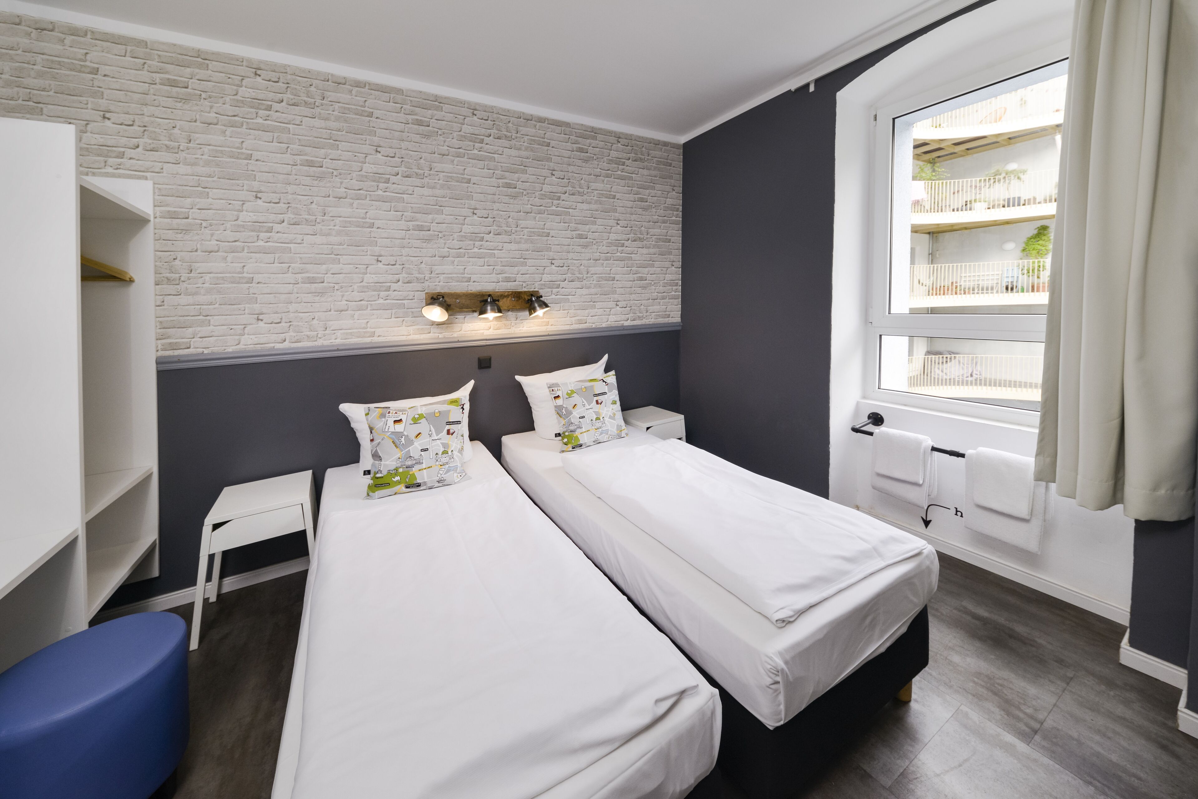 Quarto de hotel com duas camas, parede de tijolos brancos, janela com vista para varandas e uma pequena mesa branca.