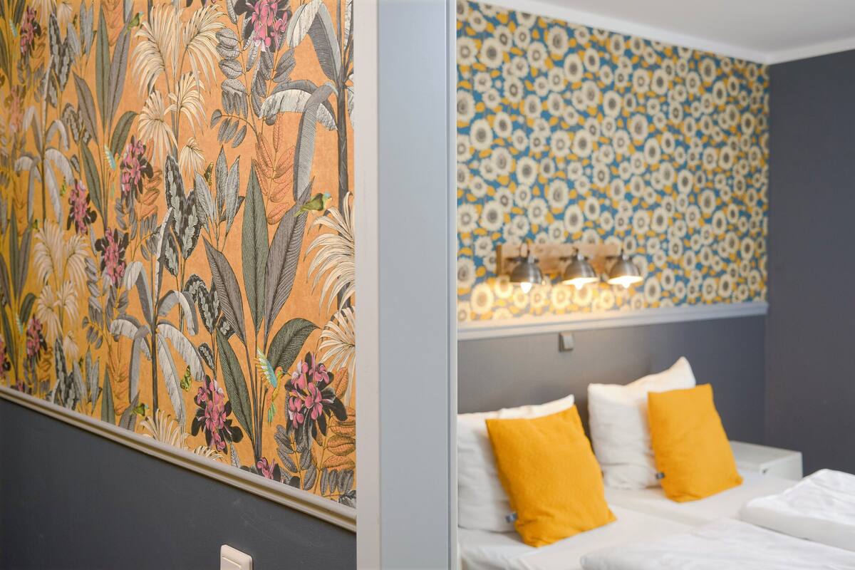 A hotel room with a bed, two yellow pillows, a floral-patterned wall, and a large floral tapestry.
