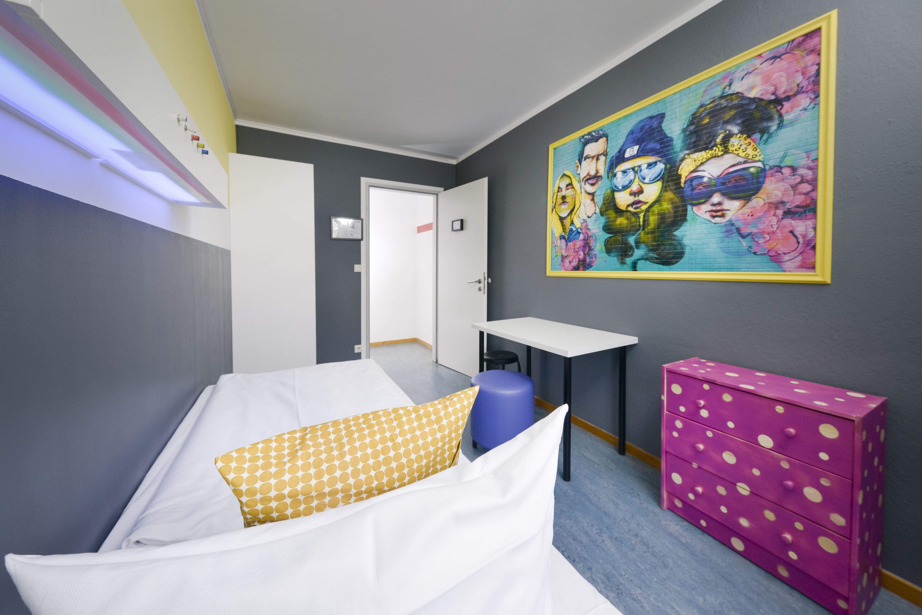 A room with a bed, a desk, a chair, a dresser, and a wall with a colorful painting.
