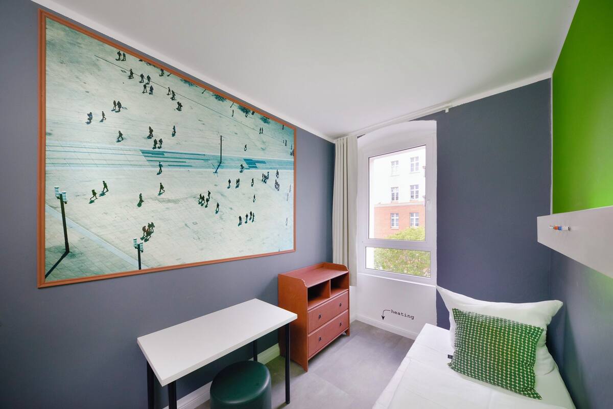 A bedroom with a large abstract painting, a desk, a dresser, a bed with a green pillow, and a window with a view of buildings.