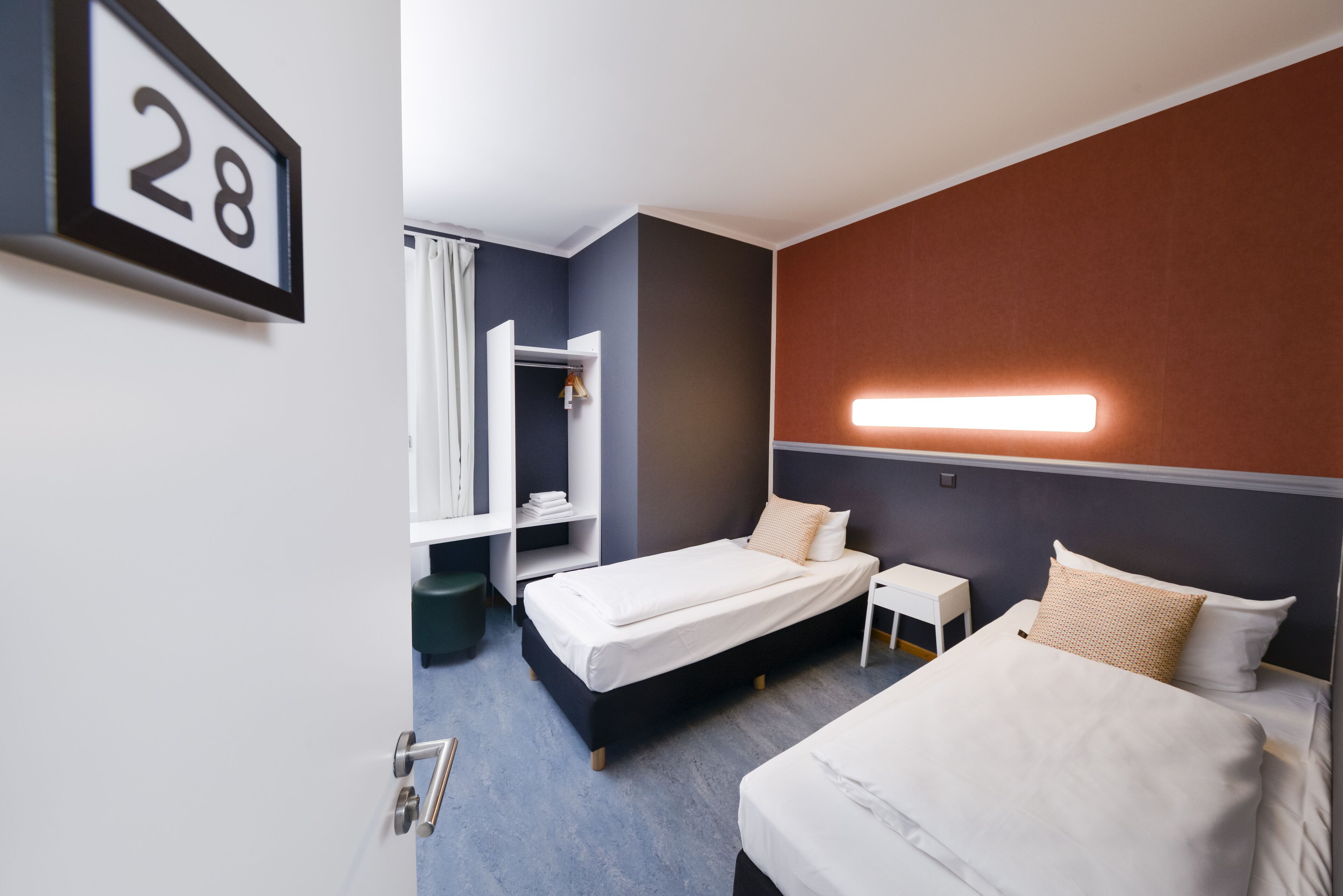 A hotel room with two beds, a wardrobe, a bedside table, and a wall-mounted light.