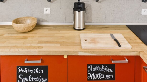 Una cocina con estantes de madera que sostienen vajilla blanca, un cuenco de madera, un hervidor y una tabla de cortar.
