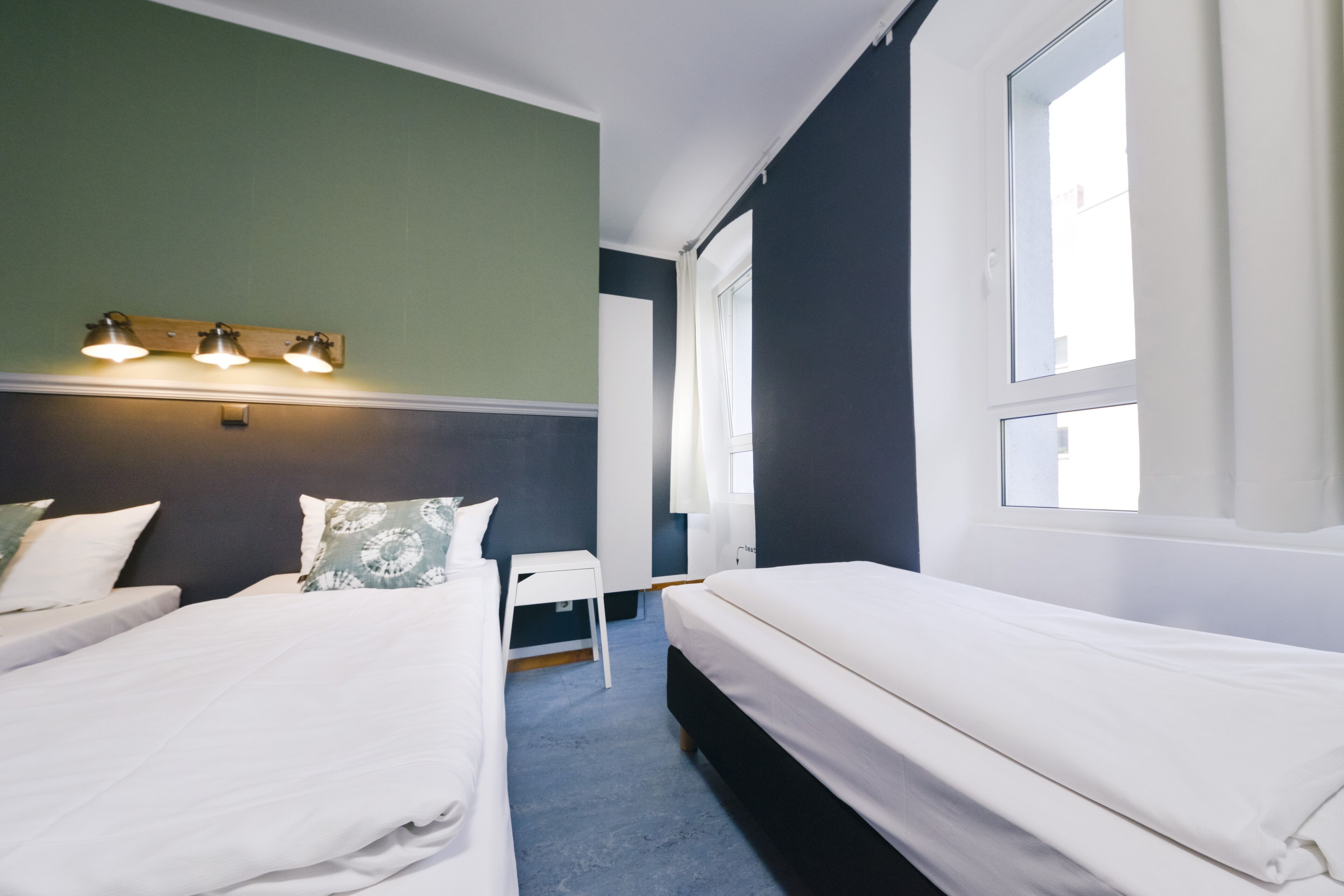 Habitación de hotel con dos camas individuales, una mesita blanca, un espejo y una ventana con cortinas.