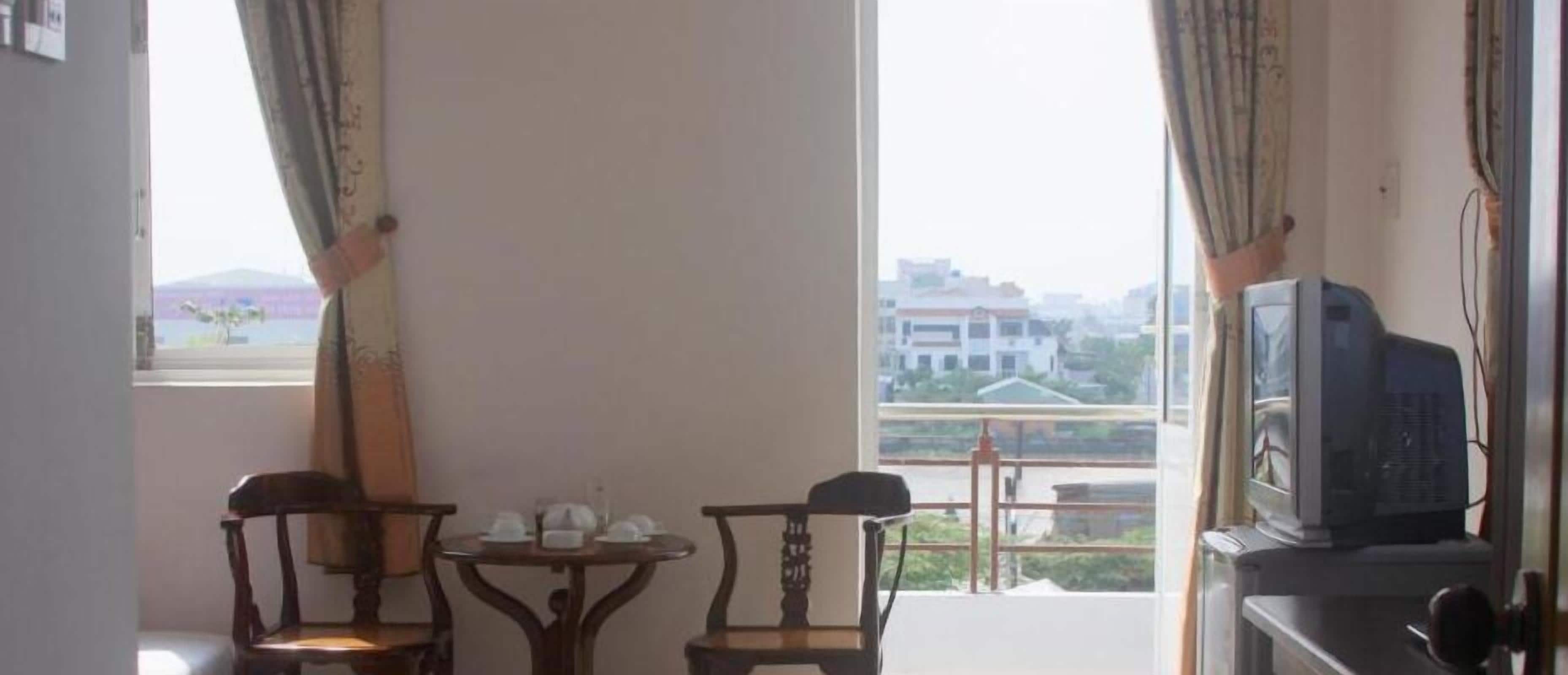 A room with two wooden chairs, a small table, a television, and a balcony with a view of buildings and a mountain.