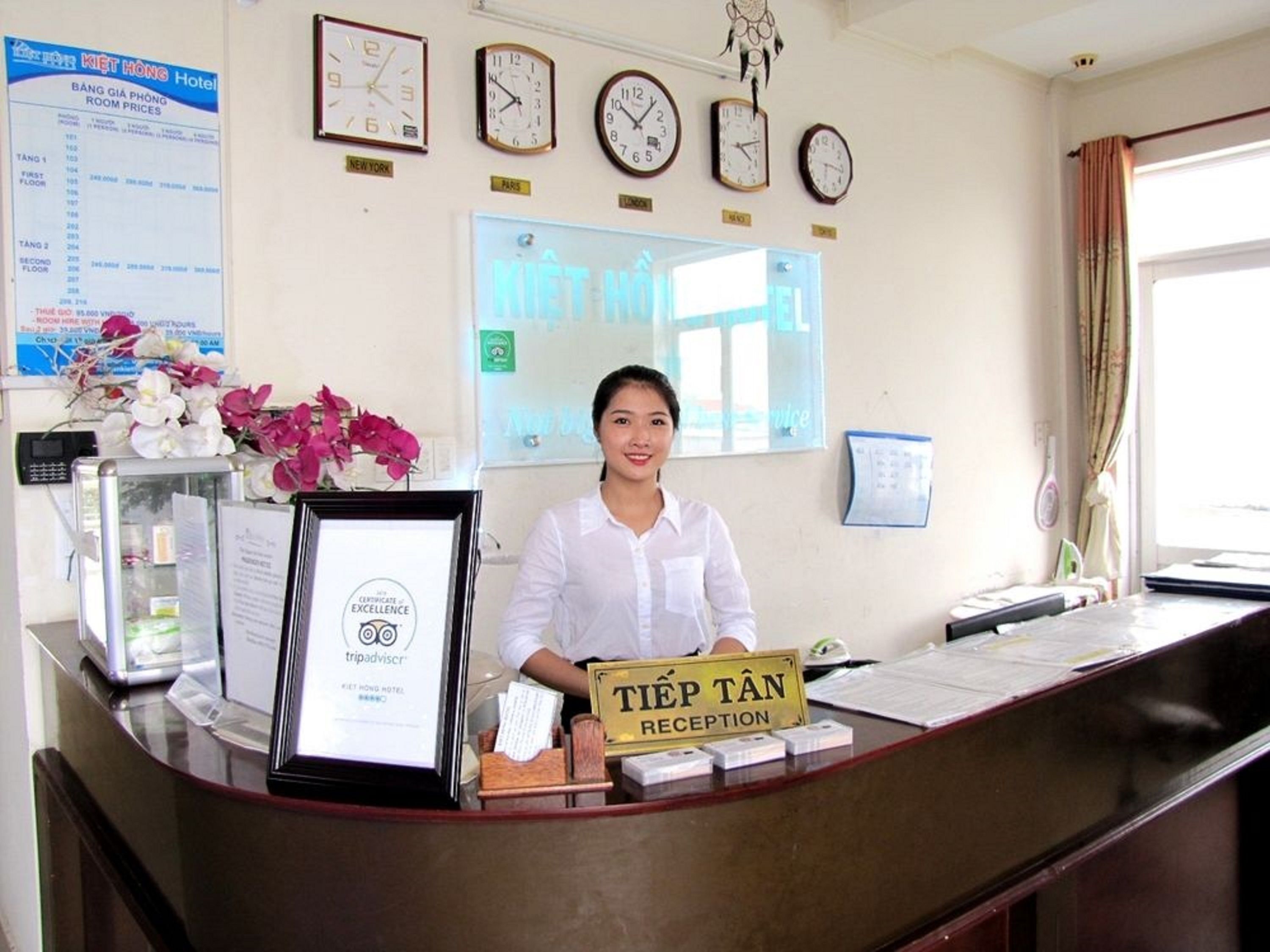 A hotel reception desk with clocks on the wall, a framed certificate, and a sign indicating the reception area.
