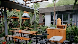 Un café al aire libre con mesas de madera y sillas negras, rodeado de exuberante follaje y una estructura de concreto.