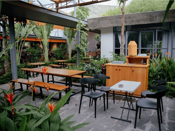 Un café al aire libre con mesas de madera y sillas negras, rodeado de exuberante follaje y una estructura de concreto.