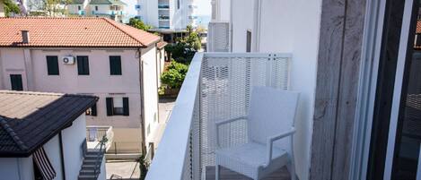 A balcony with a white wicker chair, a white railing, and a view of neighboring buildings.