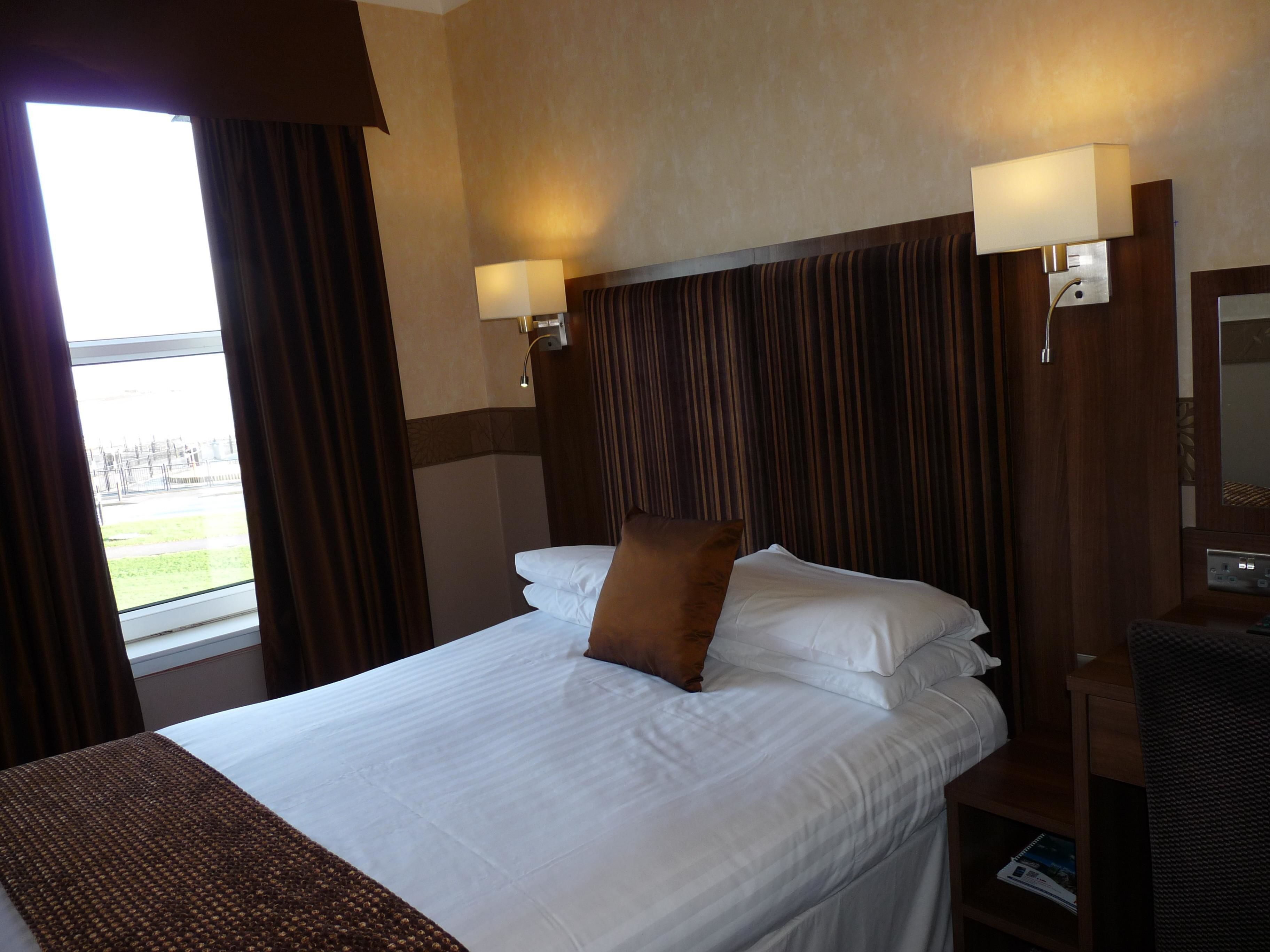 Habitación de hotel con una cama grande, dos lámparas de noche, una mesita de noche y vistas a un parque por la ventana.