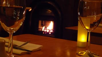 Una mesa de comedor con dos copas de vino, una vela y cubiertos. Al fondo, una chimenea con fuego encendido.