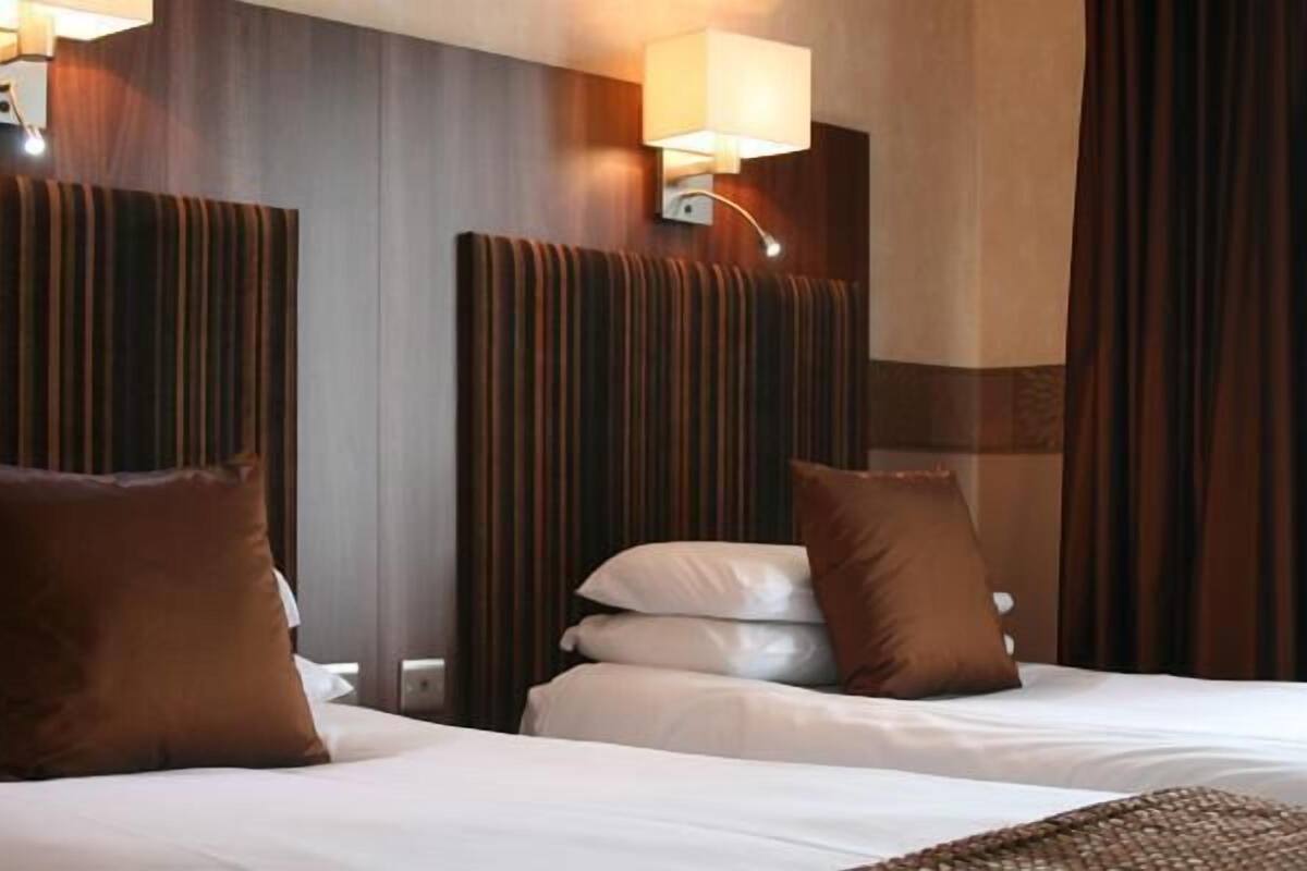 A hotel room with two beds, each with white linens and brown pillows. There are wall-mounted lamps on either side of the beds.