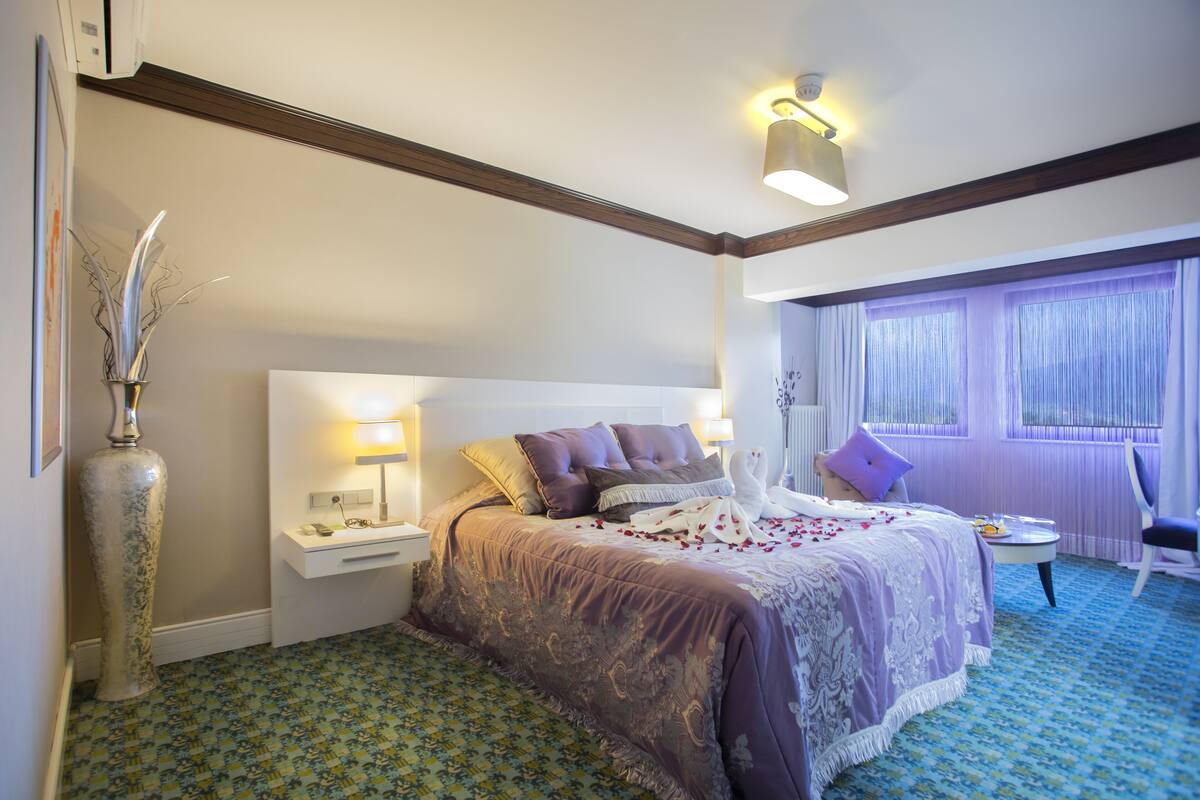 Habitación de hotel con una cama grande, una mesita de noche, un florero, una silla y una ventana con cortinas.