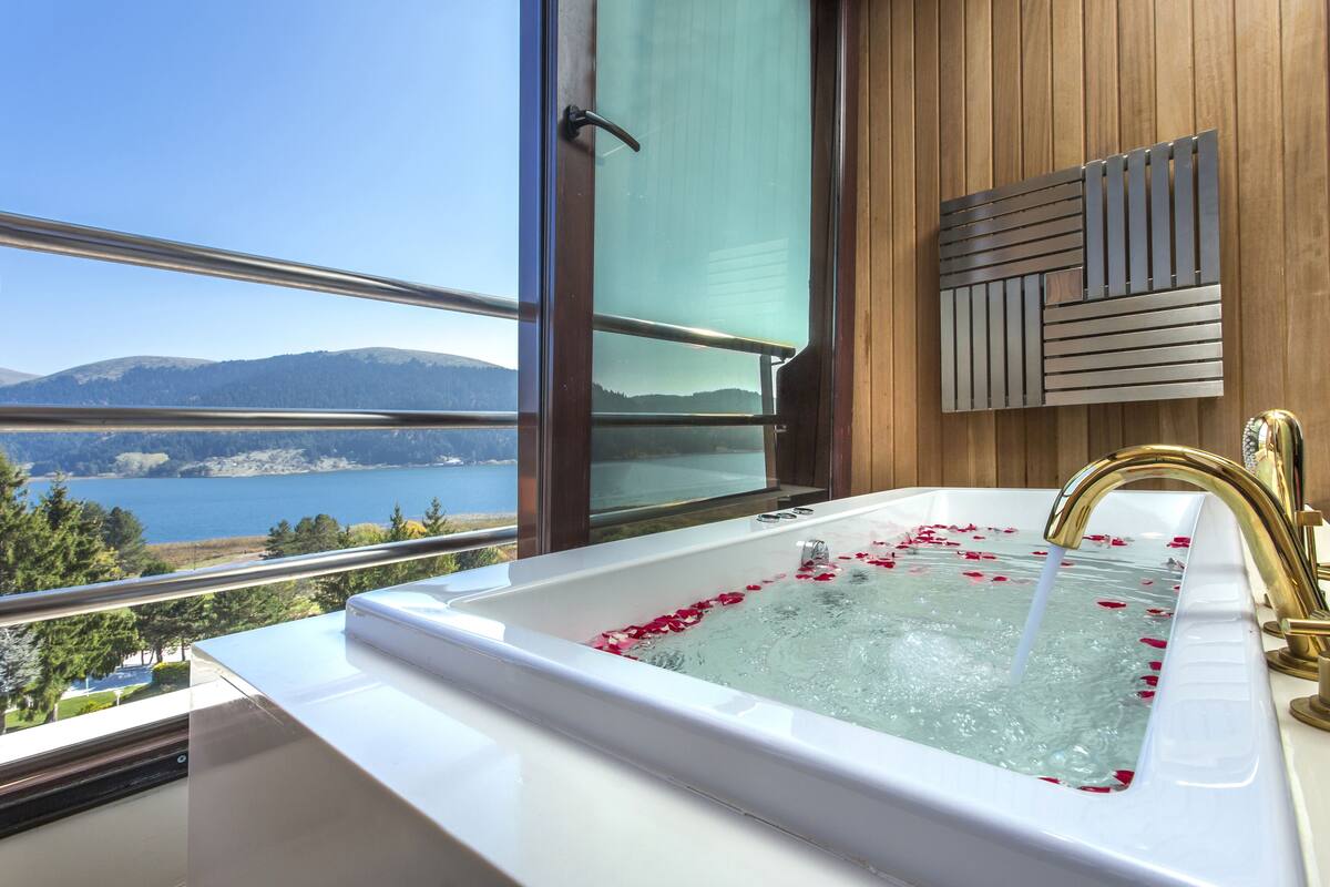 Un baño con una bañera llena de agua y pétalos de rosa, vista a un lago y montañas, y un panel de pared de madera.