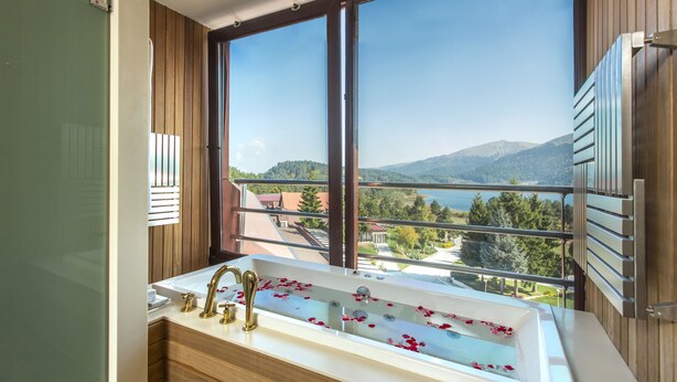 A bathroom with a large window, a bathtub with rose petals, and a view of a mountainous landscape. - Abant Palace Hotel (Mudurnu)
