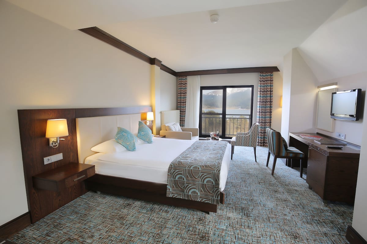 Habitación de hotel con una cama grande, un escritorio, una silla, un televisor y vista a una montaña a través de una ventana.