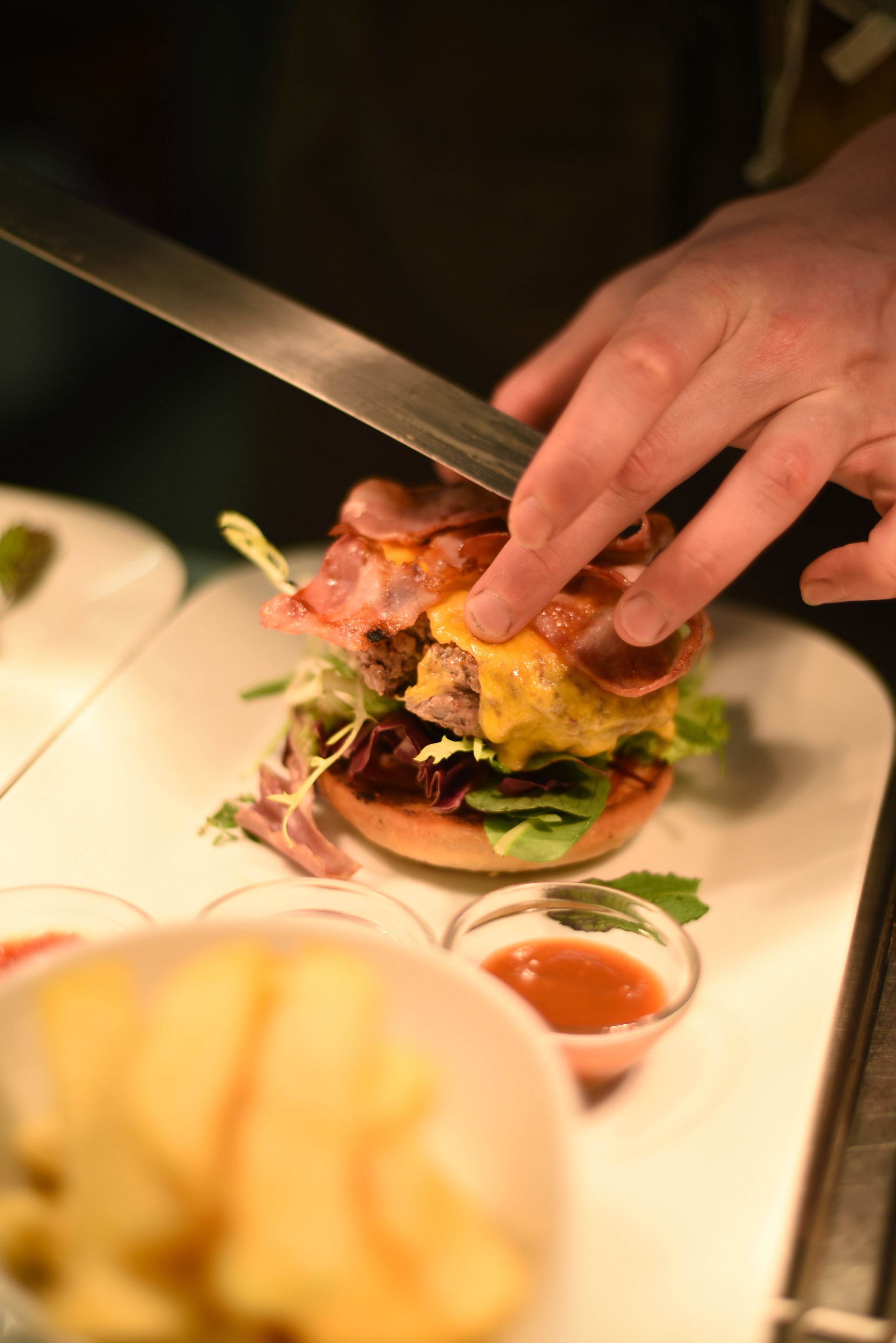 Een detailopname van een hand die een mes vasthoudt boven een hamburger met diverse toppings.