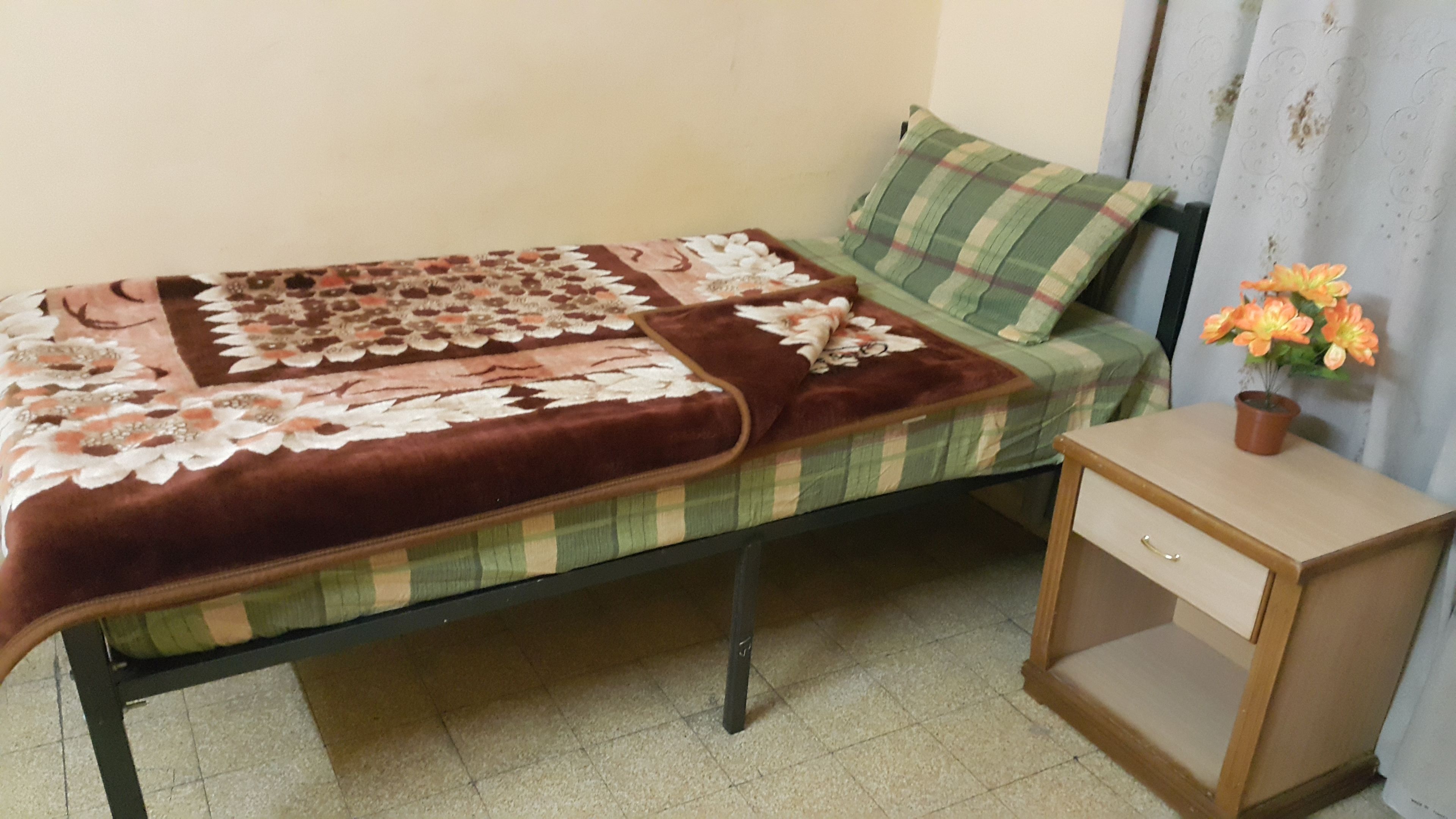 Un letto singolo con copripiumino floreale, un cuscino a quadretti verdi e beige e un comodino di legno con una pianta in vaso.