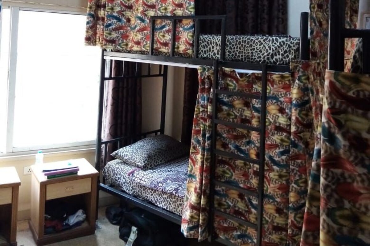 A dormitory room with bunk beds, patterned curtains, a small desk, and a window.