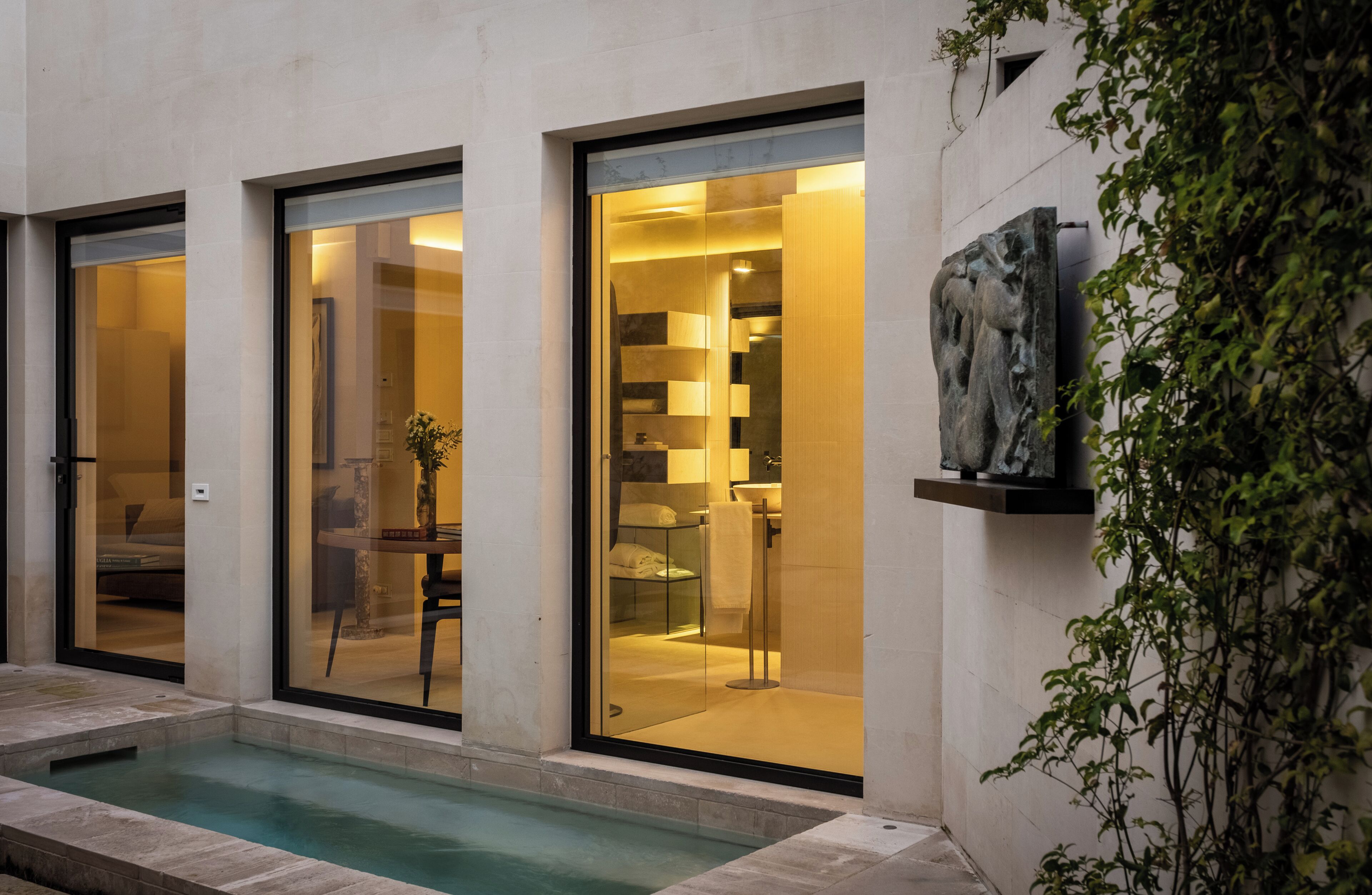 A modern building with large glass doors leading to a well-lit interior, a small pool, and a stone sculpture on the wall.