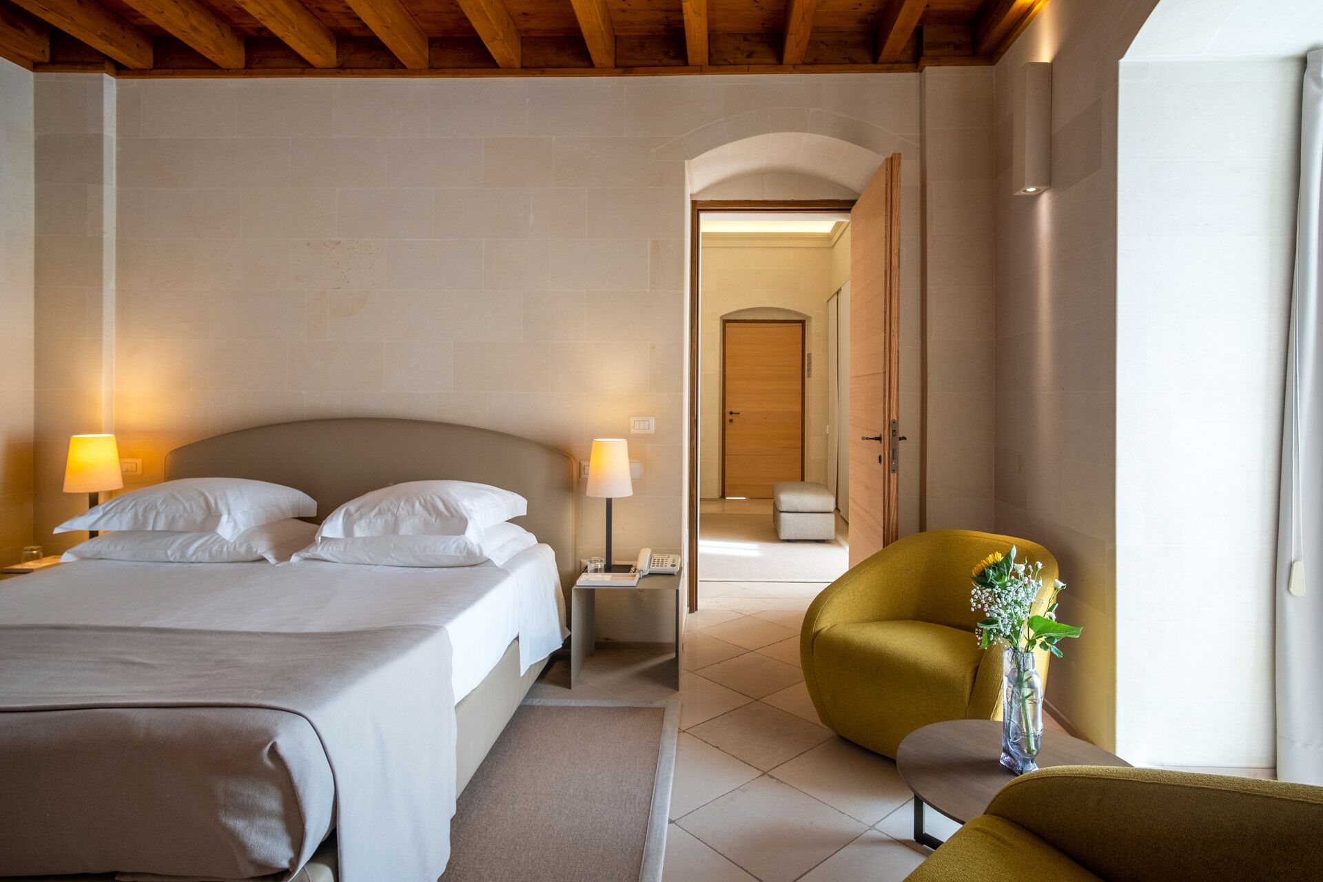 Una habitación de hotel con cama, lámparas de noche, mesita de noche, una silla y un jarrón con flores.