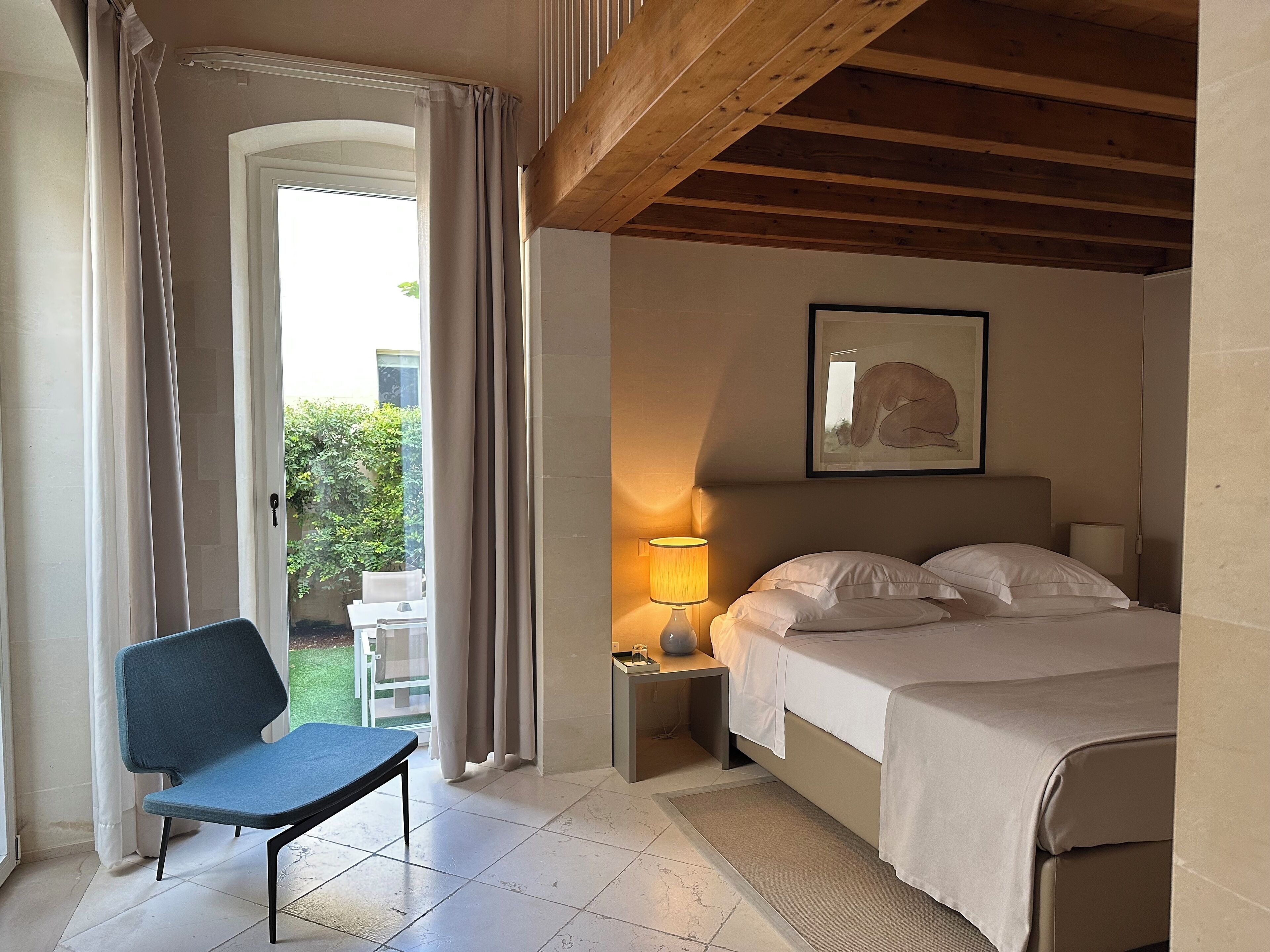 A bedroom with a bed, a chair, a nightstand with a lamp, and a painting on the wall.