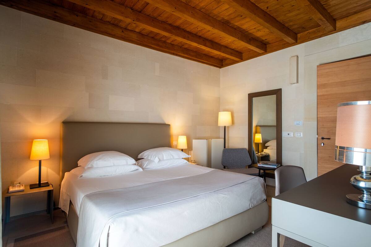 Ett hotellrum med en stor säng, två sängbord med lampor, ett skrivbord med en stol och ett träpanelat tak.