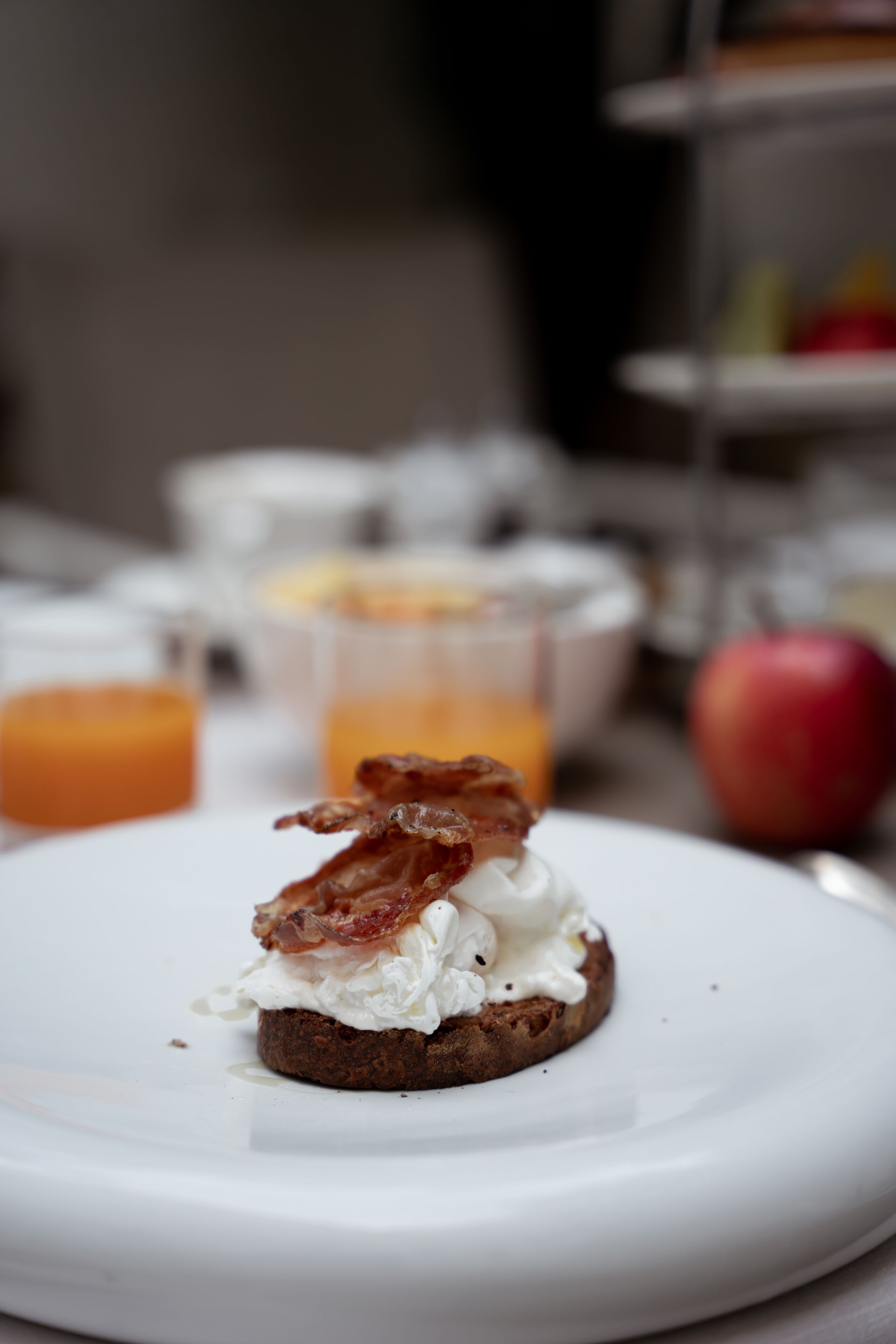 A breakfast plate with a slice of toast topped with cream and bacon, a glass of orange juice, and an apple on the side.
