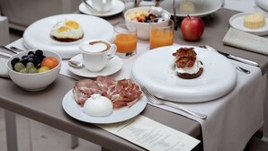 Una tavola imbandita per la colazione con piatti di cibo, tra cui uova, frutta e pasticcini, in una zona pranzo all'aperto con sedie e una pianta in vaso sullo sfondo.