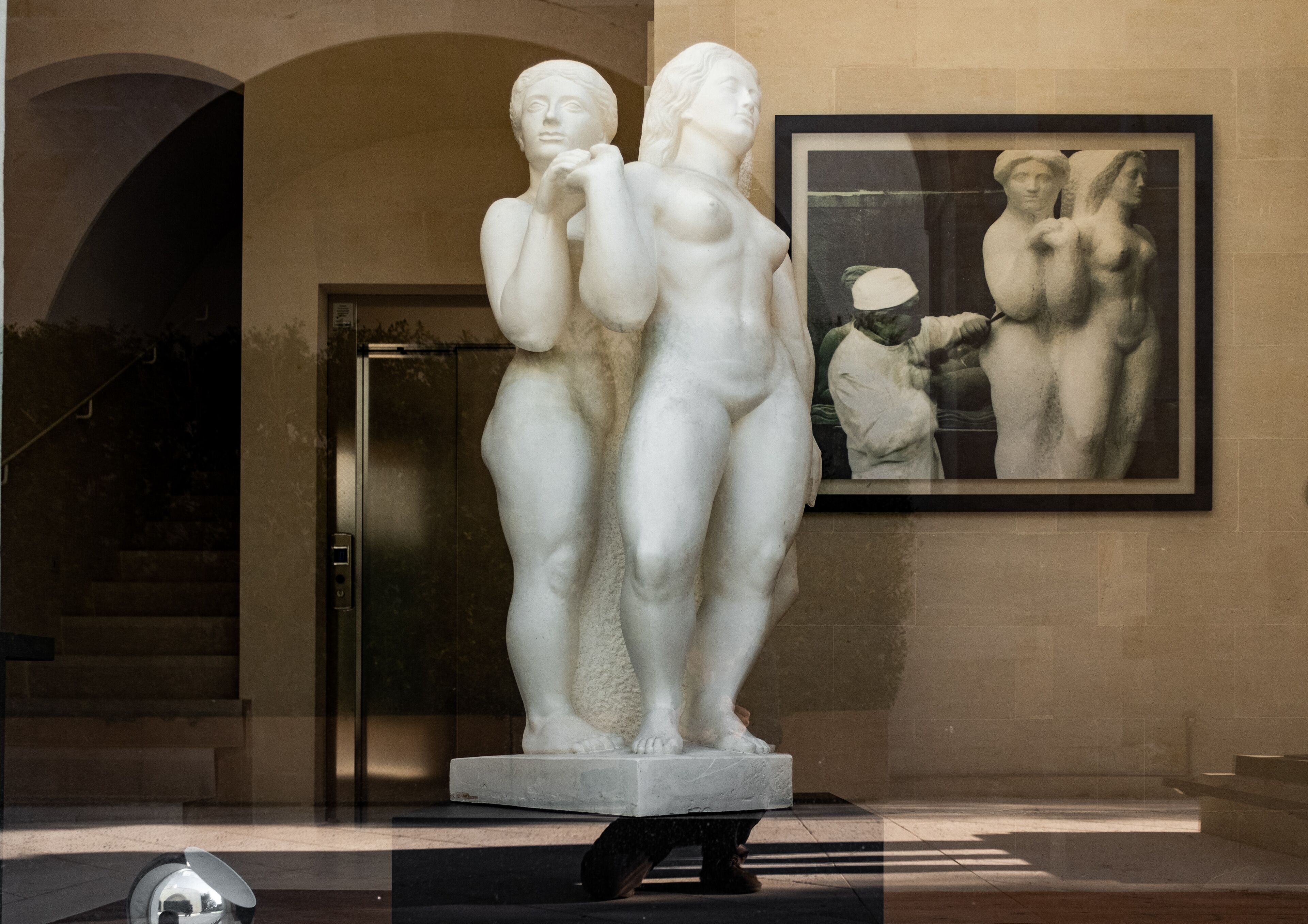 Una estatua de mármol que representa a dos figuras en un museo, con un cuadro enmarcado en la pared de fondo.