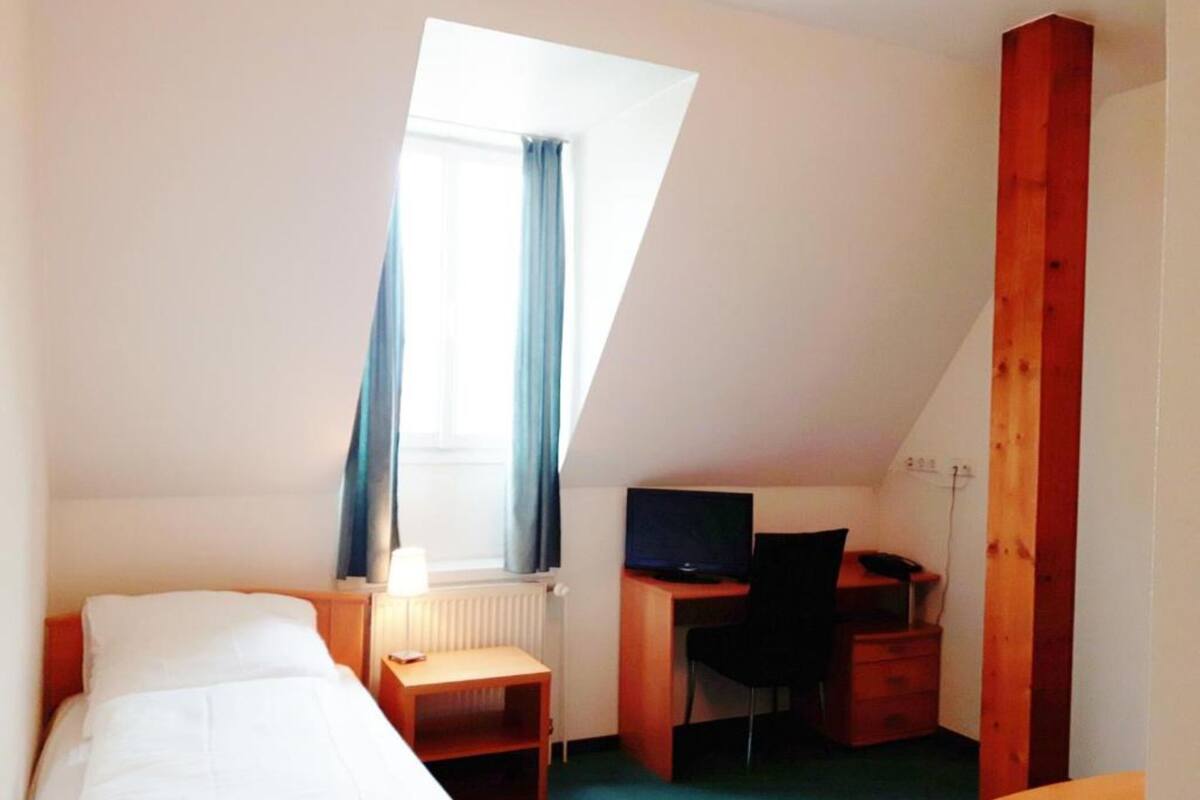 Ein kleiner, hell eingerichteter Raum mit einem Bett, einem Schreibtisch, einem Stuhl und einem Fernseher.