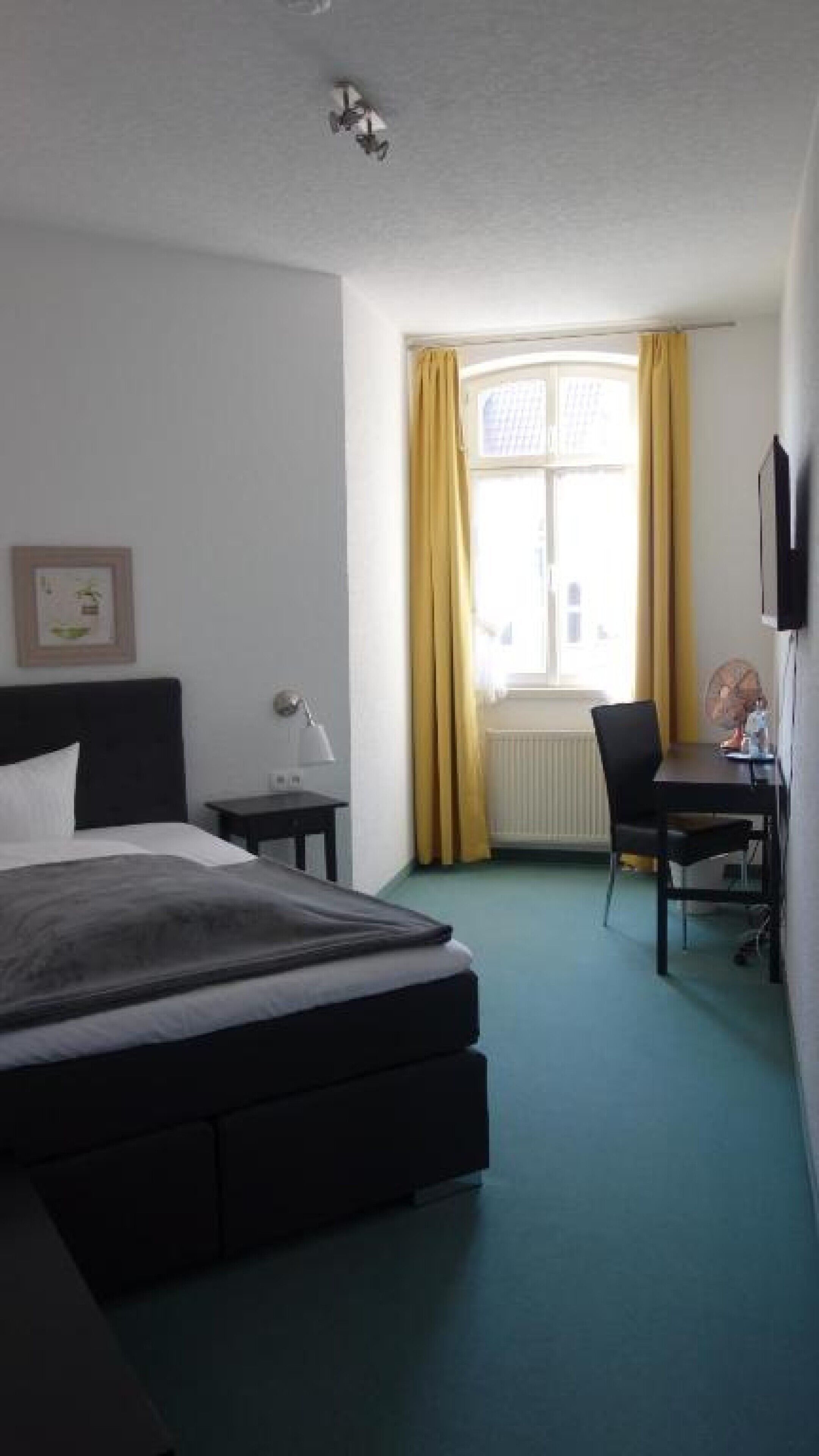 A hotel room with a bed, a desk, a chair, a radiator, and a window with curtains.