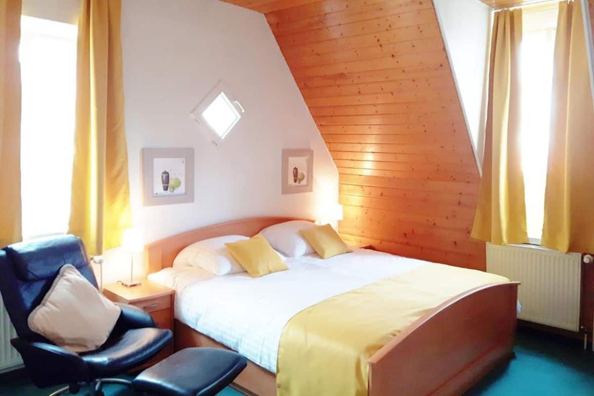 Ein Schlafzimmer mit Holzbalkendecke, einem Bett mit weißer Bettwäsche und gelben Kissen, einem schwarzen Sessel und zwei gerahmten Bildern an der Wand.