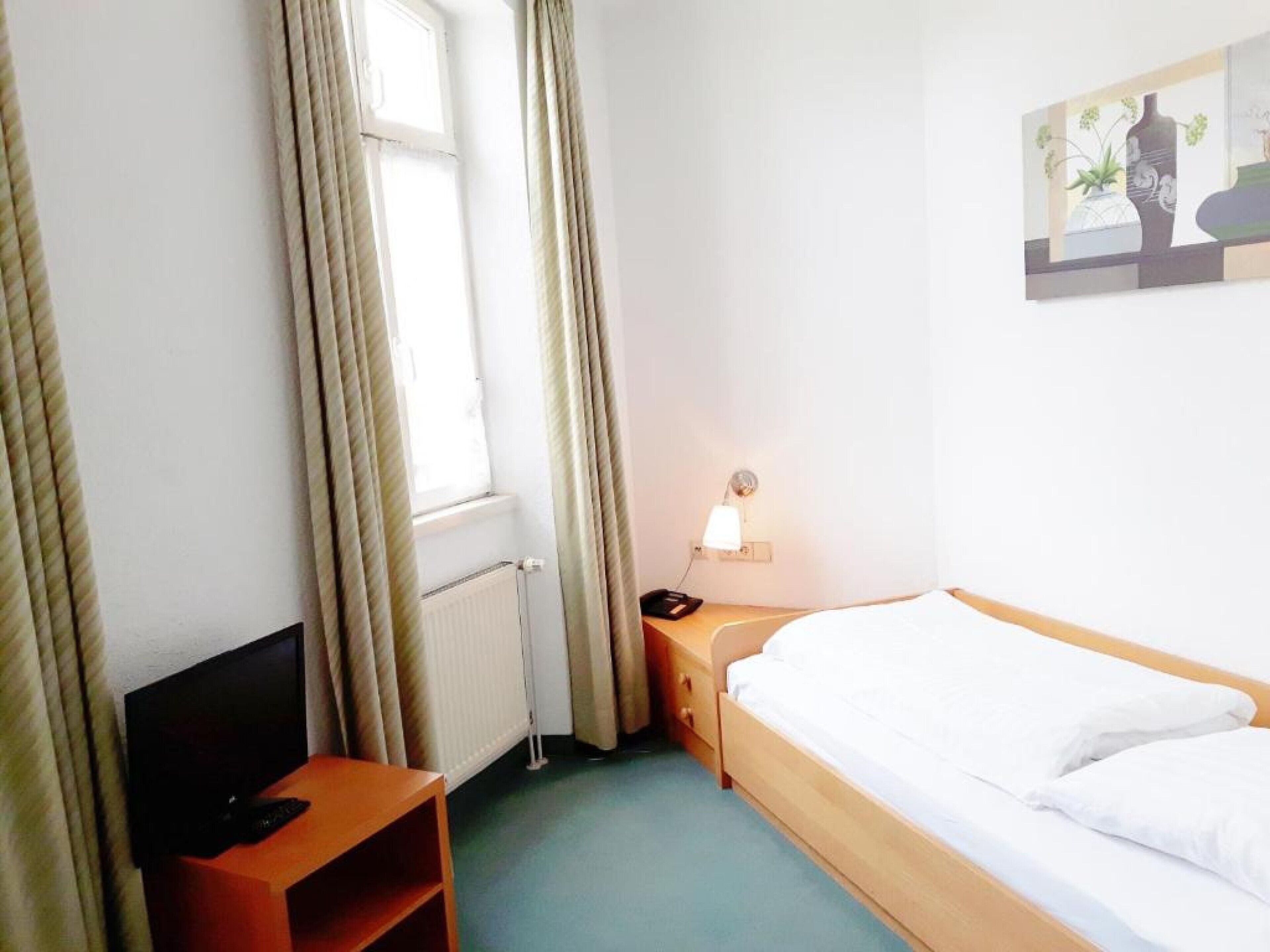 Habitación de hotel con cama, escritorio con lámpara, televisor y ventana con cortinas.