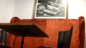 Een hoek van een kamer met een rode bekleden zitbank, een zwart-wit foto in een lijst aan de muur, en een tafel met een stoel.