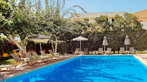 Una piscina con sillas de descanso y sombrillas, rodeada de vegetación y un edificio al fondo.