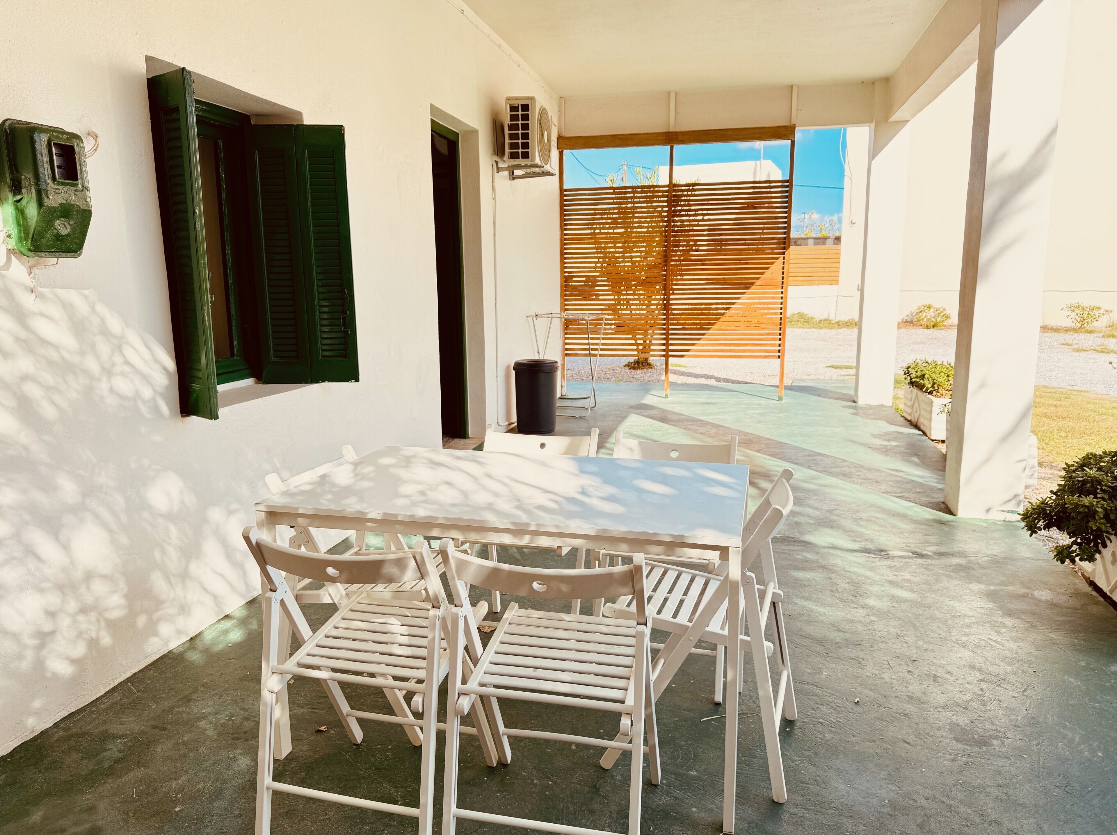 A patio with a white table and chairs, a green shuttered door, and a wooden pergola.