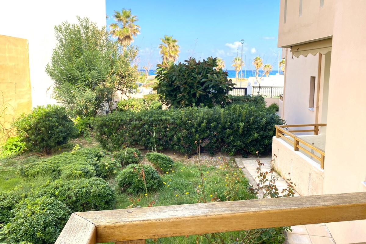 A courtyard with dense greenery, a wooden railing, and a view of the sea and palm trees in the background.