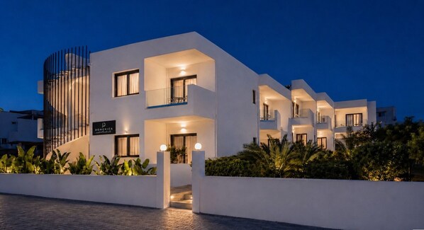 A modern hotel with a white facade, multiple balconies, and a well-lit entrance. - Domenica Apartments (Rethymno)