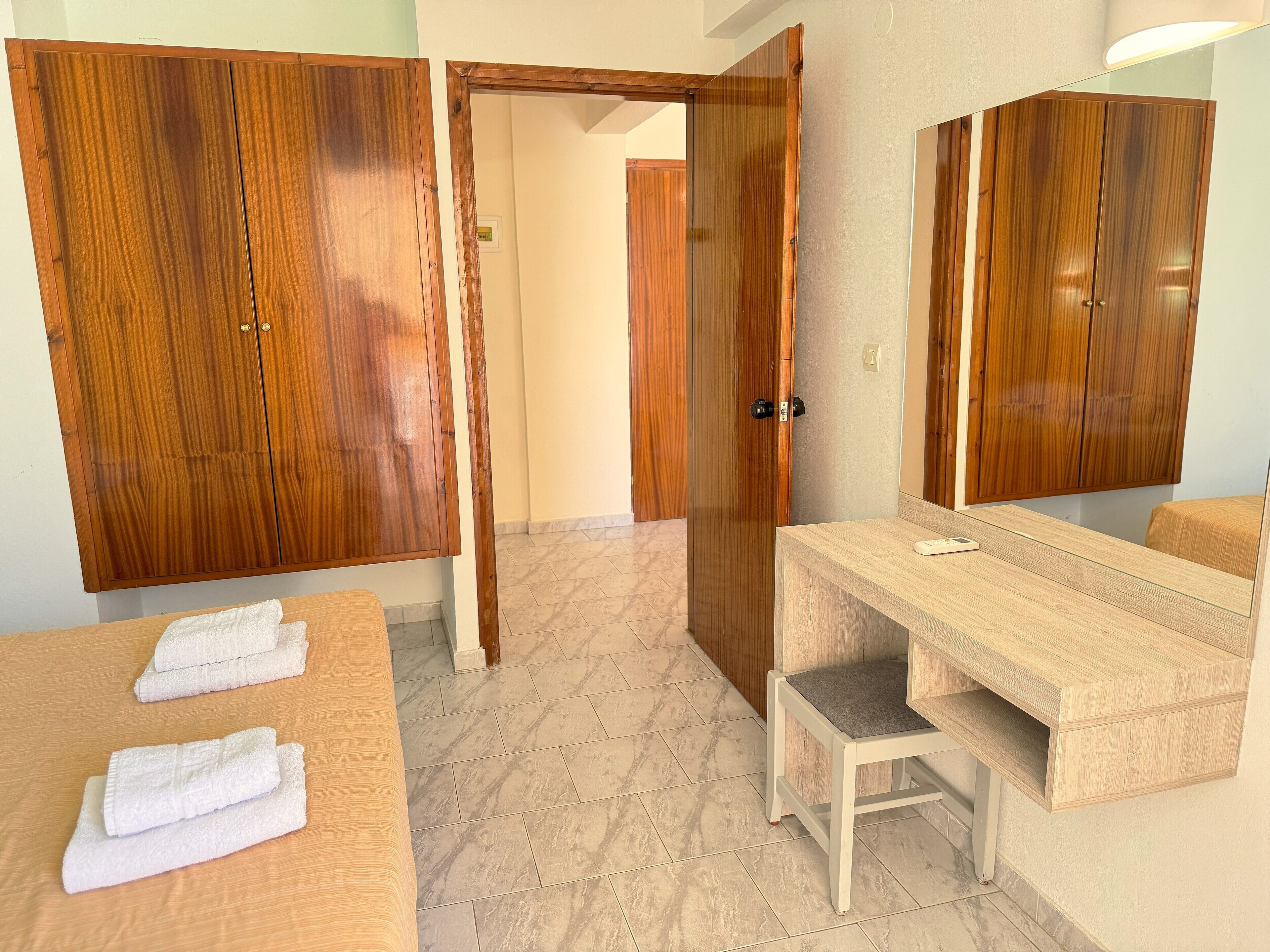 Habitación de hotel con armarios de madera, cama con toallas blancas, escritorio de madera y baño con espejo.