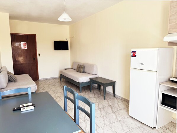 A small, well-lit room with a sofa, a table, a chair, a TV mounted on the wall, and a refrigerator. - Domenica Apartments (Rethymno)