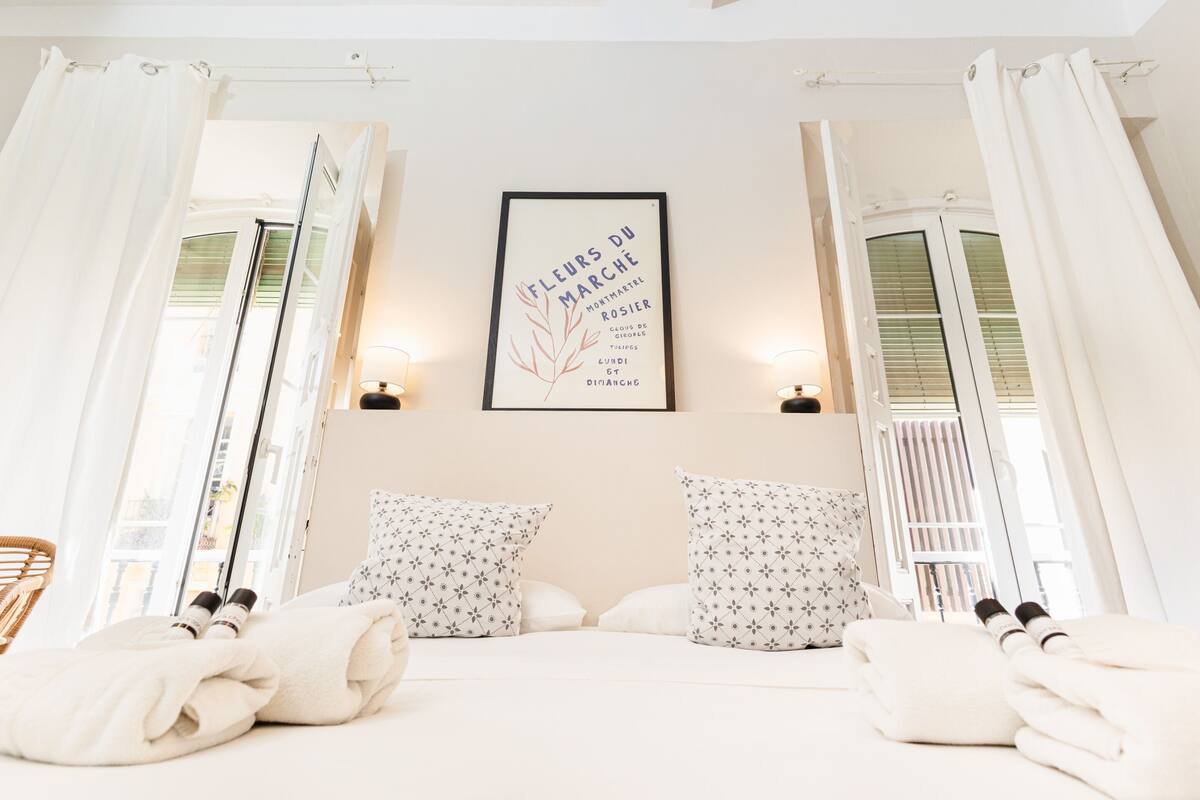 Una habitación de hotel con cama, almohadas, un cuadro en la pared y una ventana con cortinas.