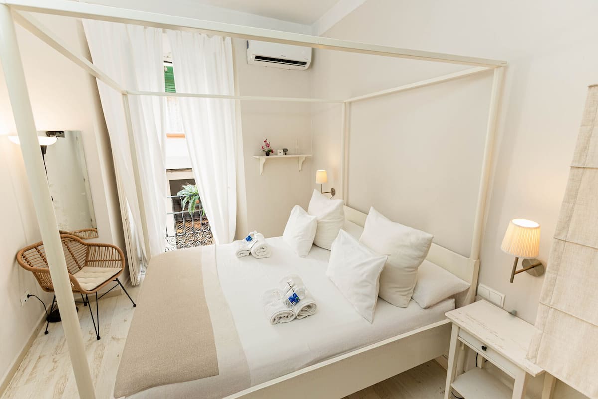 Una cama con dosel con sábanas y almohadas blancas, una mesita de noche con lámpara, una silla de mimbre y un pequeño estante con artículos de aseo.