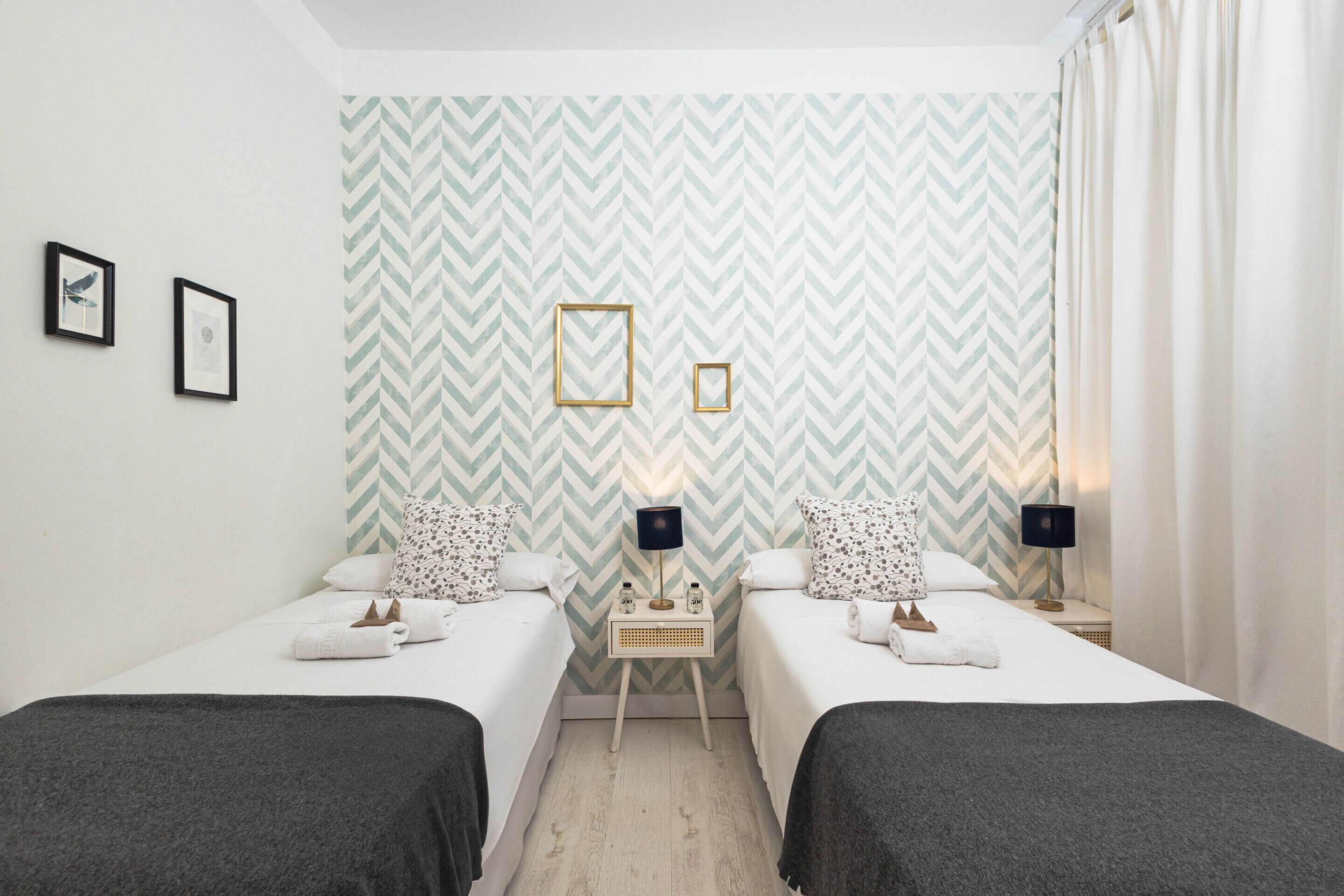 Habitación de hotel con dos camas individuales, una mesita de noche pequeña con lámpara y una pared con diseño de estampilla en zigzag.