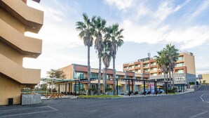 Un hotel con diseño moderno, que cuenta con una zona de estar al aire libre cubierta, palmeras y un estacionamiento.