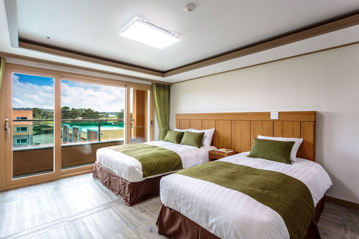 A hotel room with two beds, a large window with a view of a pool and buildings, and a ceiling light.