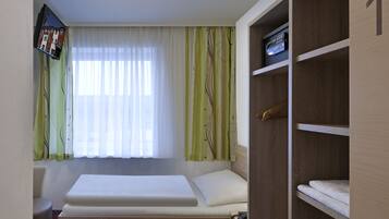 A compact room with a bed, a wooden wardrobe, a small desk, and a window with sheer curtains.