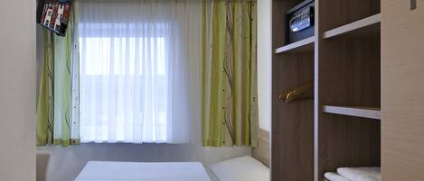 Habitación pequeña con cama, armario de madera, escritorio y ventana con cortinas traslúcidas.
