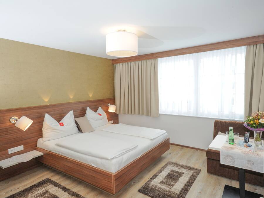 Habitación de hotel con cama de madera, lámparas de noche, una mesita con un florero y una ventana con cortinas.