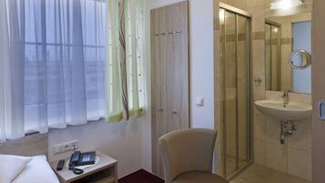 A hotel room with a bed, a chair, a small table with a phone, a window with sheer curtains, and a bathroom with a sink and mirror.