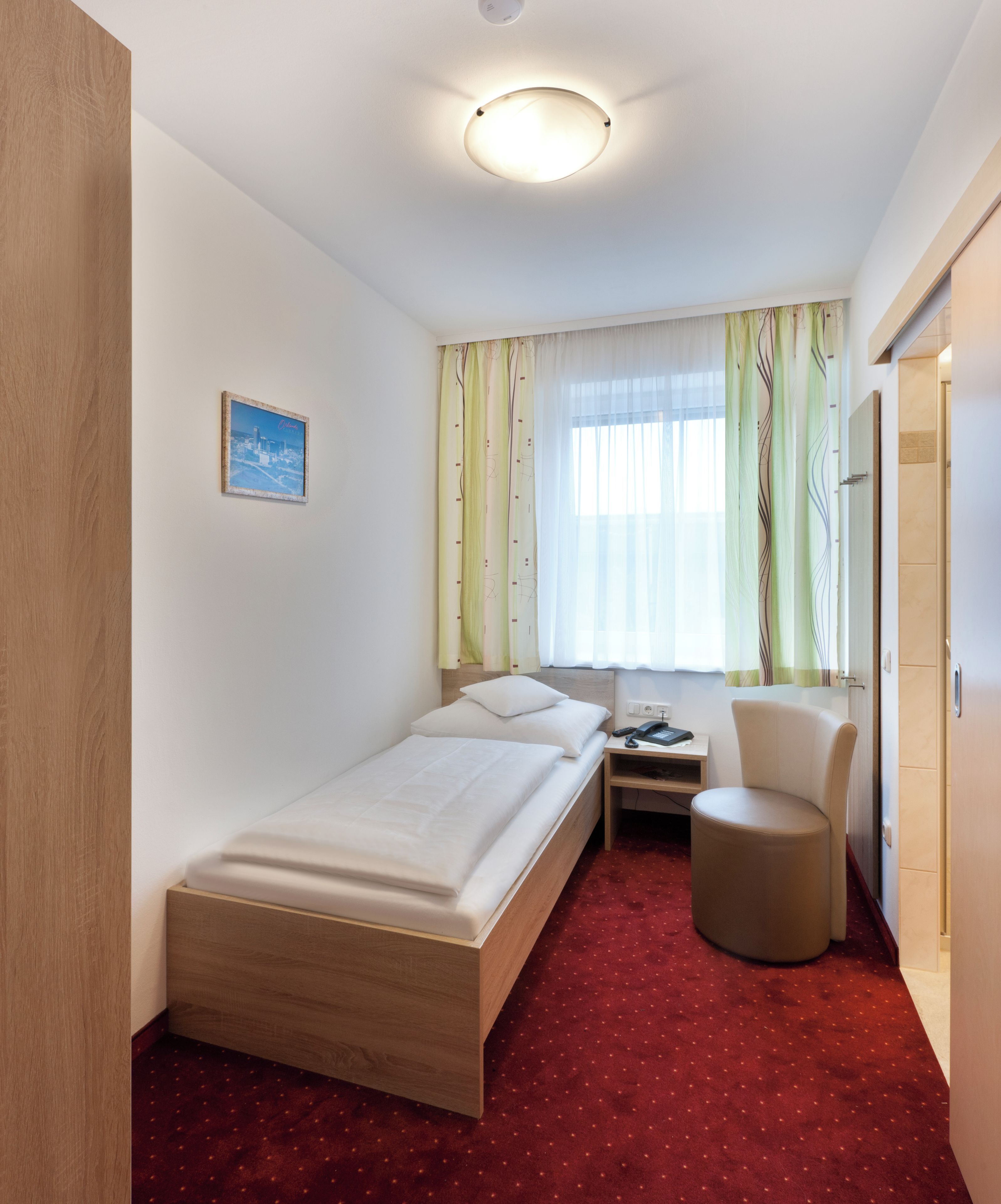Habitación de hotel con una cama individual, un escritorio, una silla y un otoman redondo pequeño.